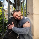 Two adult brothers enjoy a warm embrace in a sunny park. They stand beside a decorative fence surrounded by lush greenery, showcasing a strong bond and happiness in the moment.