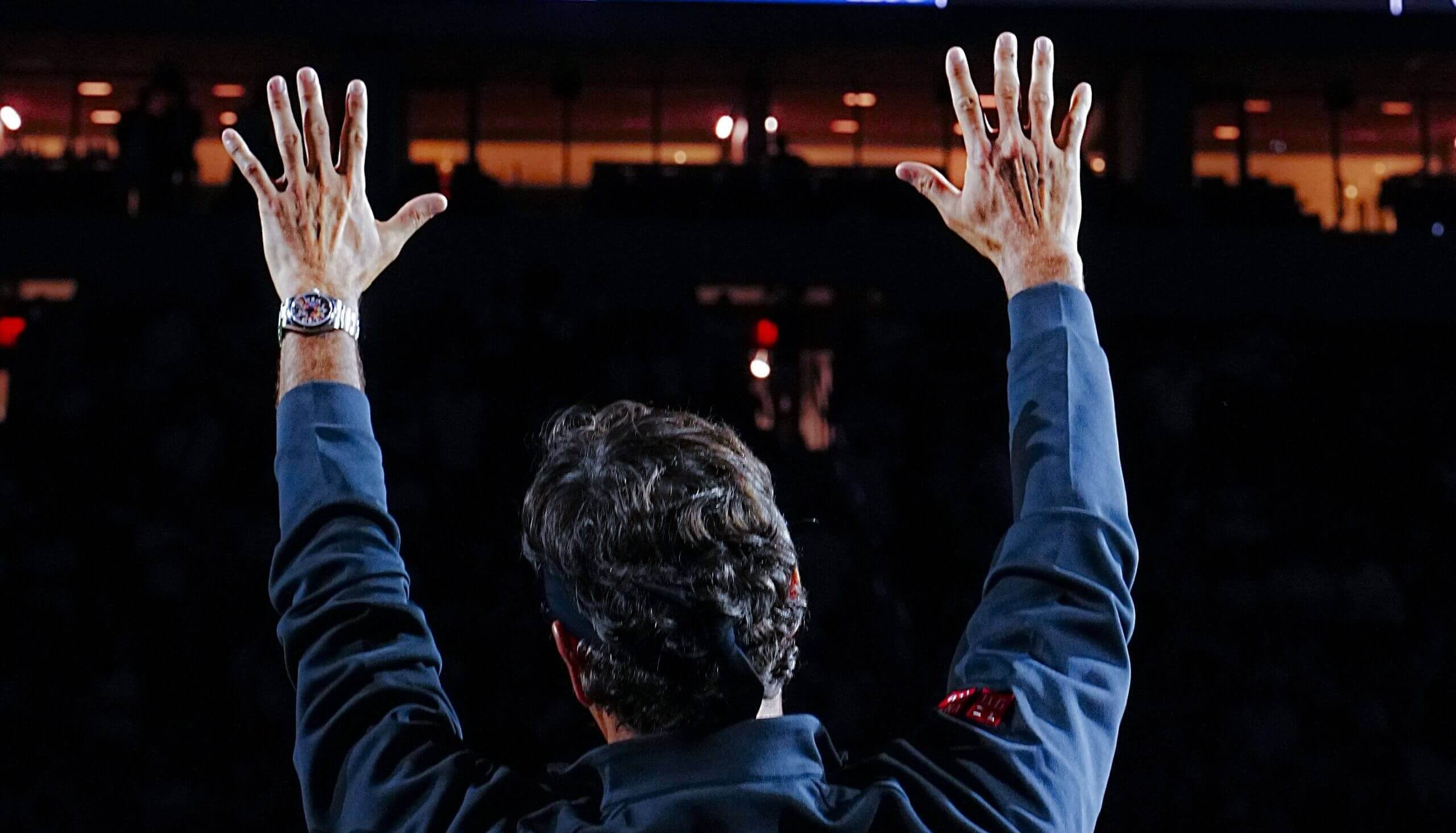 Roger Federer raises his hands to acknowledge a crowd unseen because of darkness. The camera is behind him.