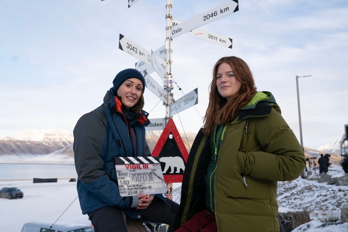 Vigil_SuranneJones_Rose Leslie Suranne Jones and Rose Leslie on the set of Vigil season 3 wearing puffer coats and holding a clapperboard.