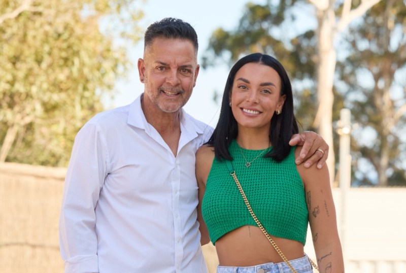 Two contestants from "The Block" pose together outdoors, with trees in the background. The man wears a white shirt and smiles confidently, while the woman sports a green crop top and denim shorts, showcasing a cheerful expression.