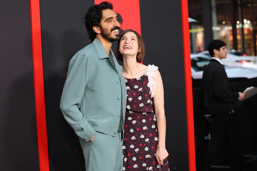 woman wears a patterned dress on a red carpet and looks up at her partner who wears light blue suit. Red and black backdrop