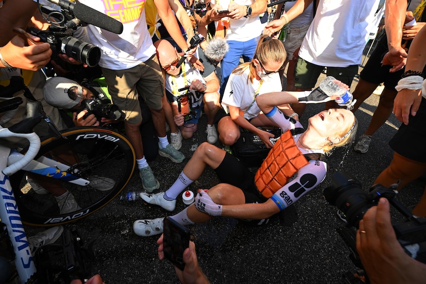 An exhausted cyclist pours water on her head while sitting on the ground.
