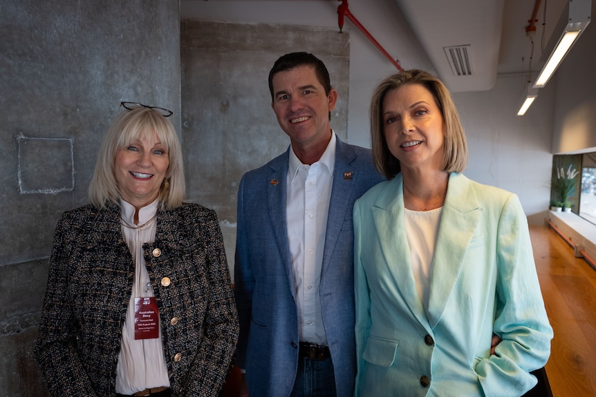 A woman with blonde hair stands next to a man in a light blue suit and a woman in a pastel green suit jacket, all smiling.