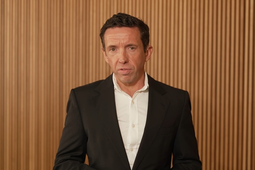 Stephen Beckett speaking seriously, wearing a black suit and white shirt and speaking in front of a wooden wall.