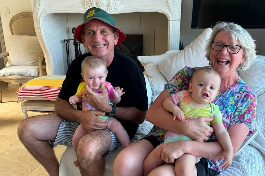 A woman and a man sit on a couch with young babies on their laps, everyone smiling for the camera