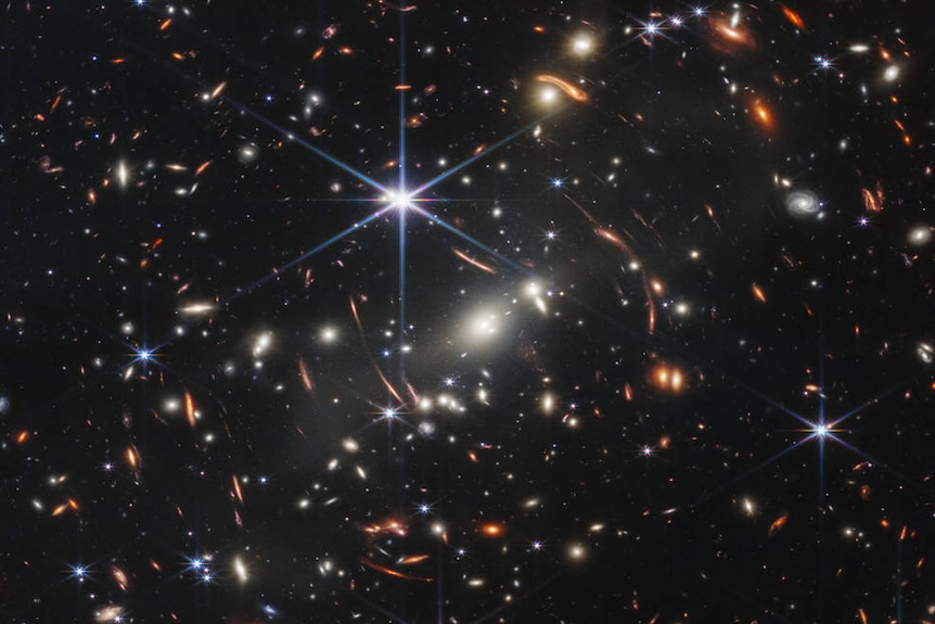 Starry field with white patch of galaxies at centre and orange-gold stretched and distorted galaxies circling it.