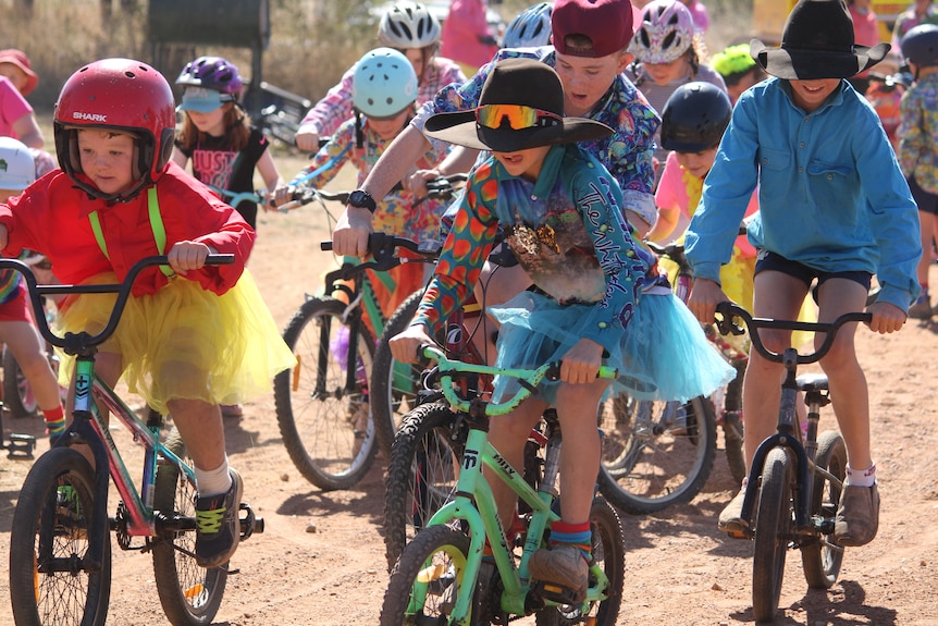 Kids and adults dressed in bright colours riding bikes and walking along a road in the outback.
