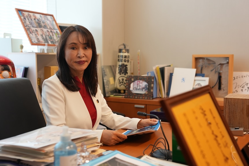 A woman in a white suit jacket and red shirt sits in an office.