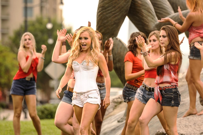 A still from Nashville. Hayden is in a park, dressed in a white corset top and shorts, and surrounded by female dancers.