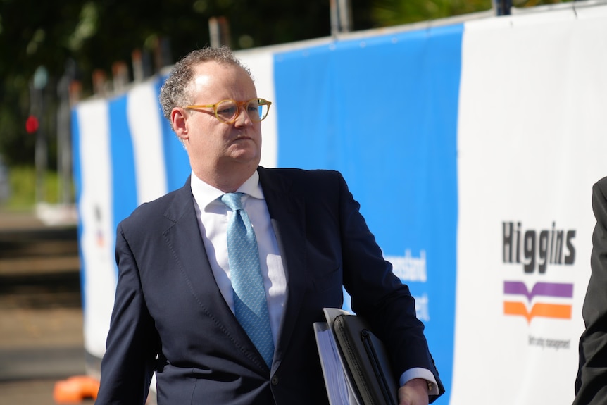 A man in a suit and tie and glasses walking while carrying documents.
