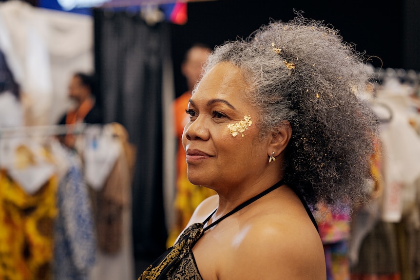 Close up shoulder shot of Mum of Natisha Tabua, who has gray curly hair with gold sparkles on her cheekbone, wearing black top.