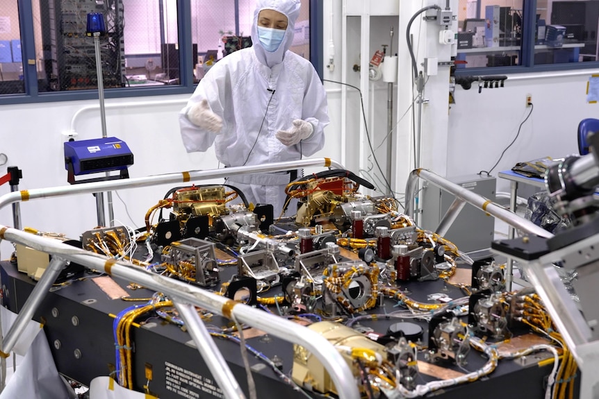 Person in a forensic suit and face mask standing behind a wedge-shaped block mounted with complicated devices and cables.