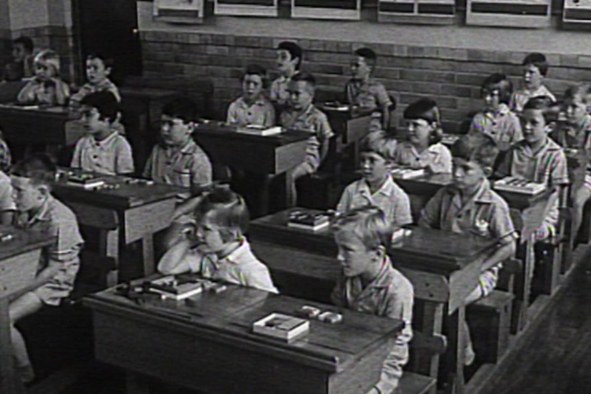 A black and white image of a classroom full of young students sitting at old wooden desks in the 1960s