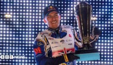A male wearing a speedway biker outfit holding a large silver trophy while smiling. He is standing in front of a wall covered in small lights.