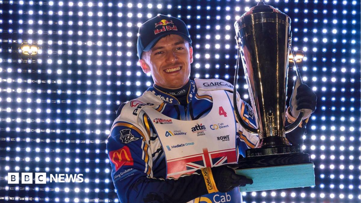 A male wearing a speedway biker outfit holding a large silver trophy while smiling. He is standing in front of a wall covered in small lights.
