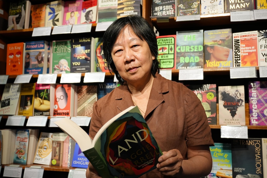 A woman, Jaye Chin-Dusting, looks serious at the camera, reading a book in front of a bookshelf