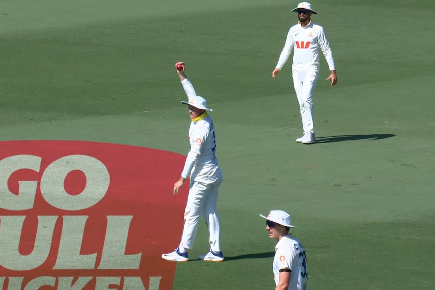 A cricketer in white holds up a ball in appeal