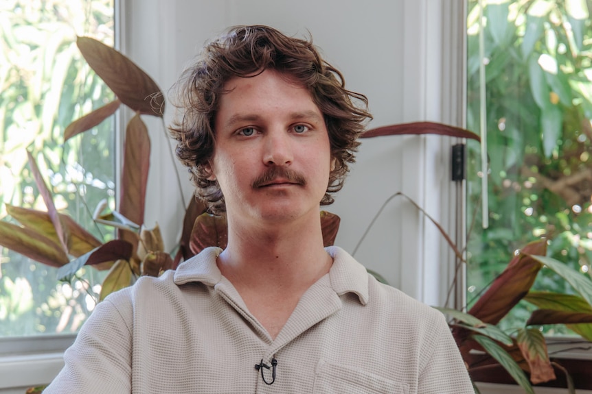 A man looks directly at the camera, sitting with one leg resting on the other.