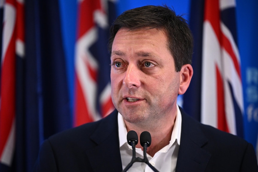 A dark-haired man stands in front of the Australian flag and speaks into a microphone. He has a neutral expression.