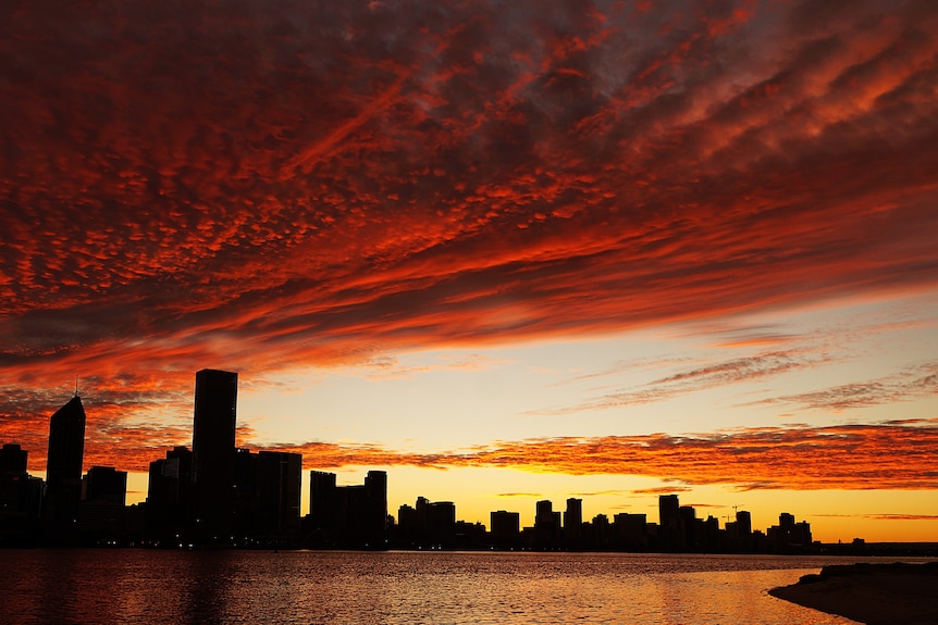 A sunrise over Perth's CBD colours the sky orange and red as clouds pass over the city, leaving clear sky.