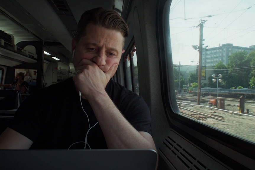 Ben McKenzie looking at a laptop critically while on a train.