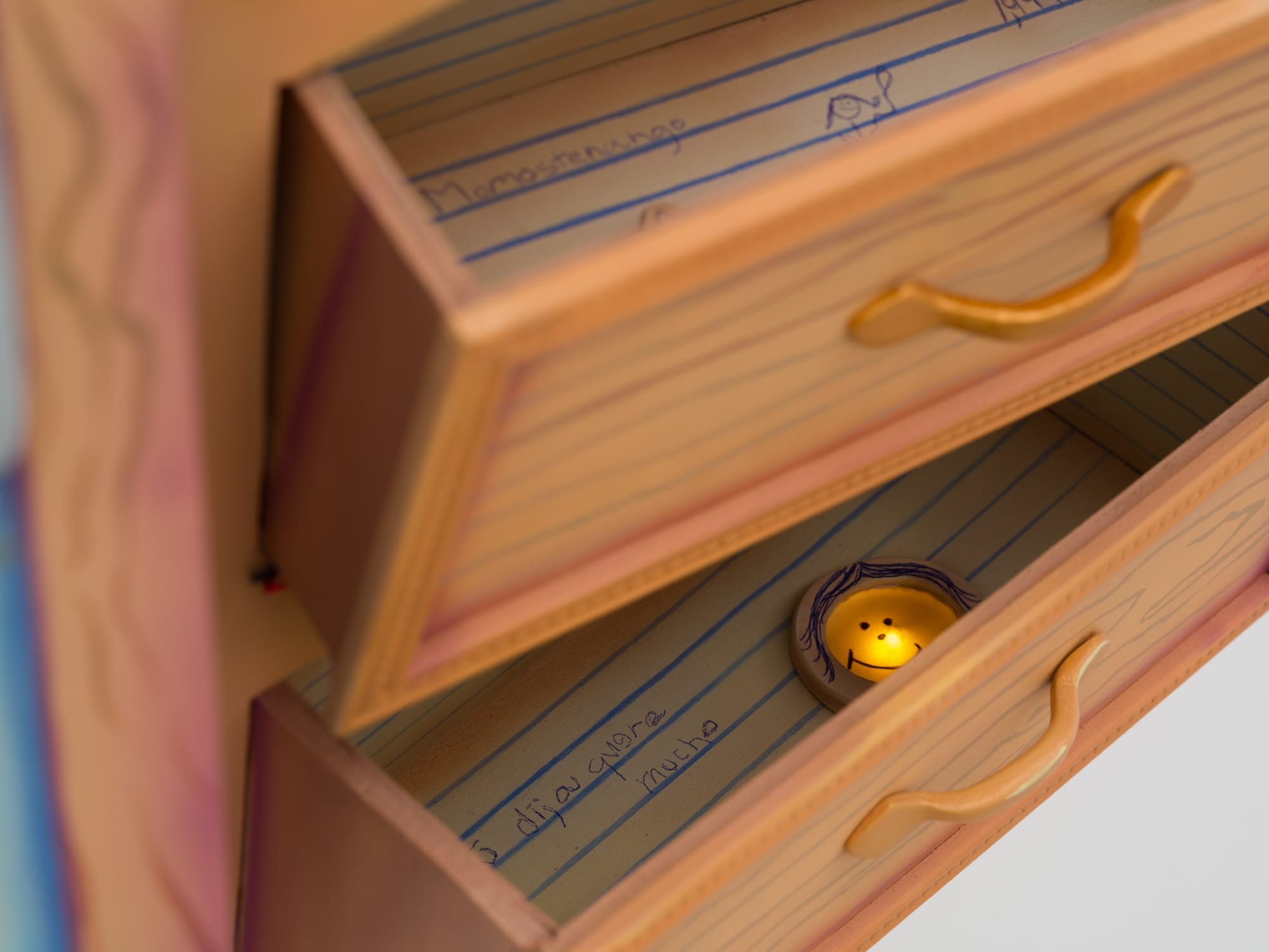 a sculptural painting by Estefania Ajcip of drawers pulled out of a dresser with lined paper inside and a smiley face light