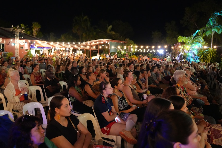 A large crowd of people looking at a stage, a string of fairy lights is twinkling in the background.