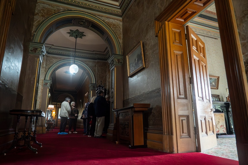 A small group gather in a wide ornate hallway of a 19th century mansion.