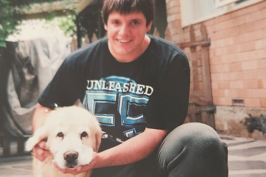 A man with short brown hair wearing a black tshirt is kneeling down and patting a dog.