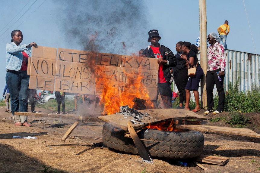 Young men and women holding a sign in front of a burning tyre on the street.