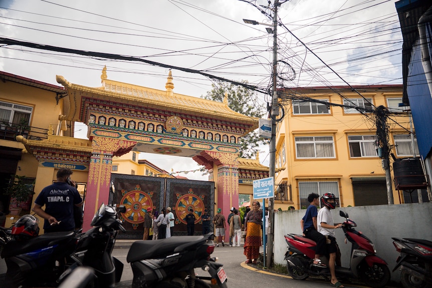 The yellow walls and ornate gates to a monastery are closed, as people stand in the street and on ride on mopeds.