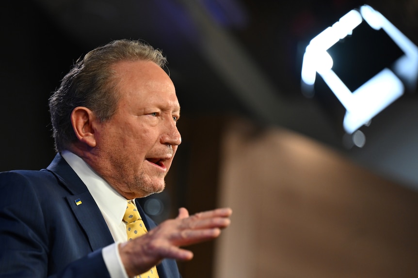 A side-on photo of a man wearing a blue suit, white shirt and yellow tie gesturing with his right hand while speaking on stage.