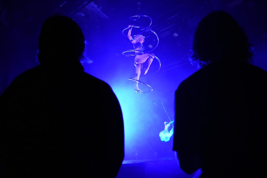 An acrobatic woman performs on an aparatus suspended from the ceiling of a darkened room