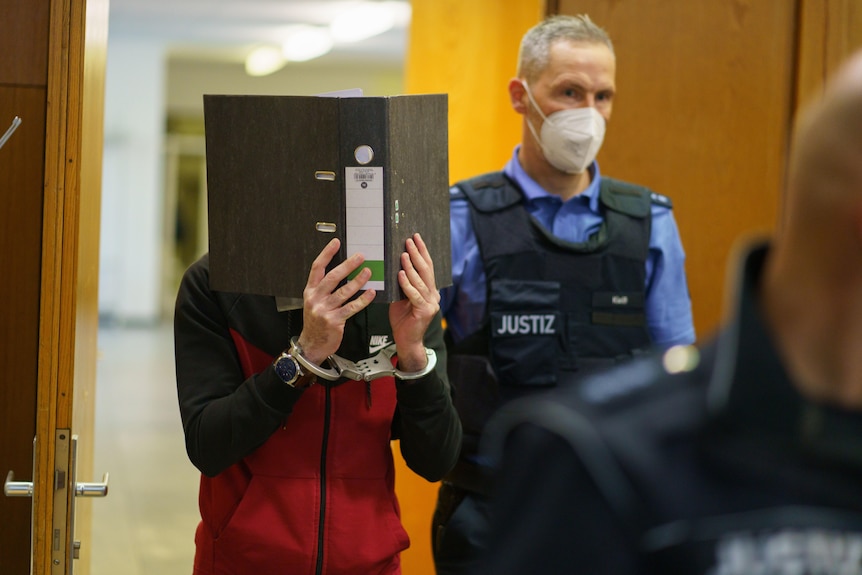 A man holds up a binder to cover his face while entering a court room.
