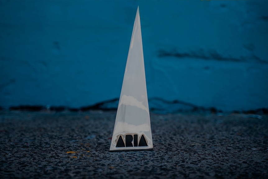 Silver pointy ARIA Award trophy emblazoned with ARIA logo, sitting on concrete against blue brick background.