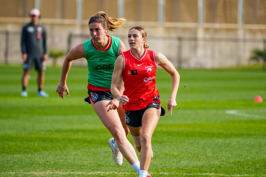 Two women one in green jersey one in red jersey running on a football field.