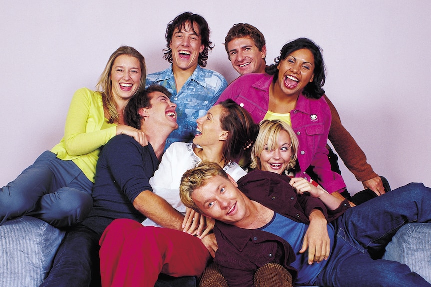 A publicity shot of a smiling group of young actors sitting together.