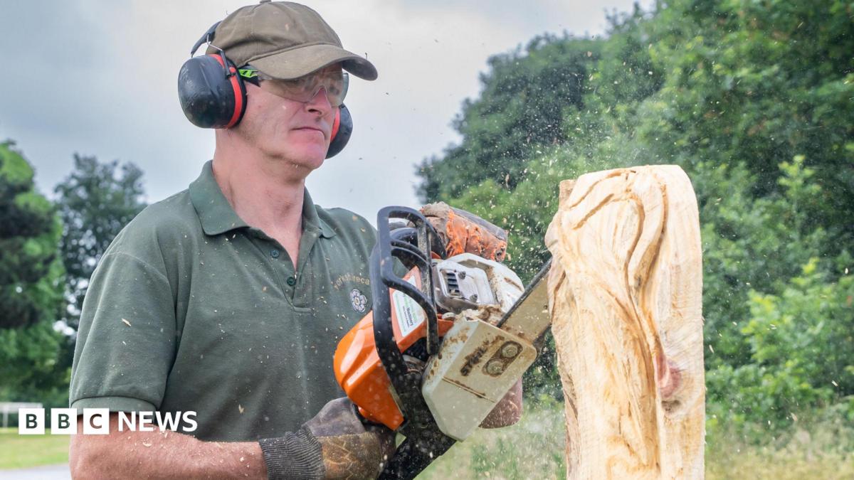 A man with a green polo shirt, cap and ear defenders holds a chainsaw as he carves a wooden sculpture.