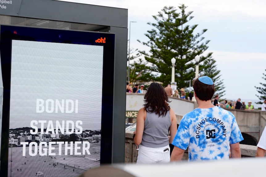 Two people walking past an electronic sign reading 'Bondi Stands Together'.