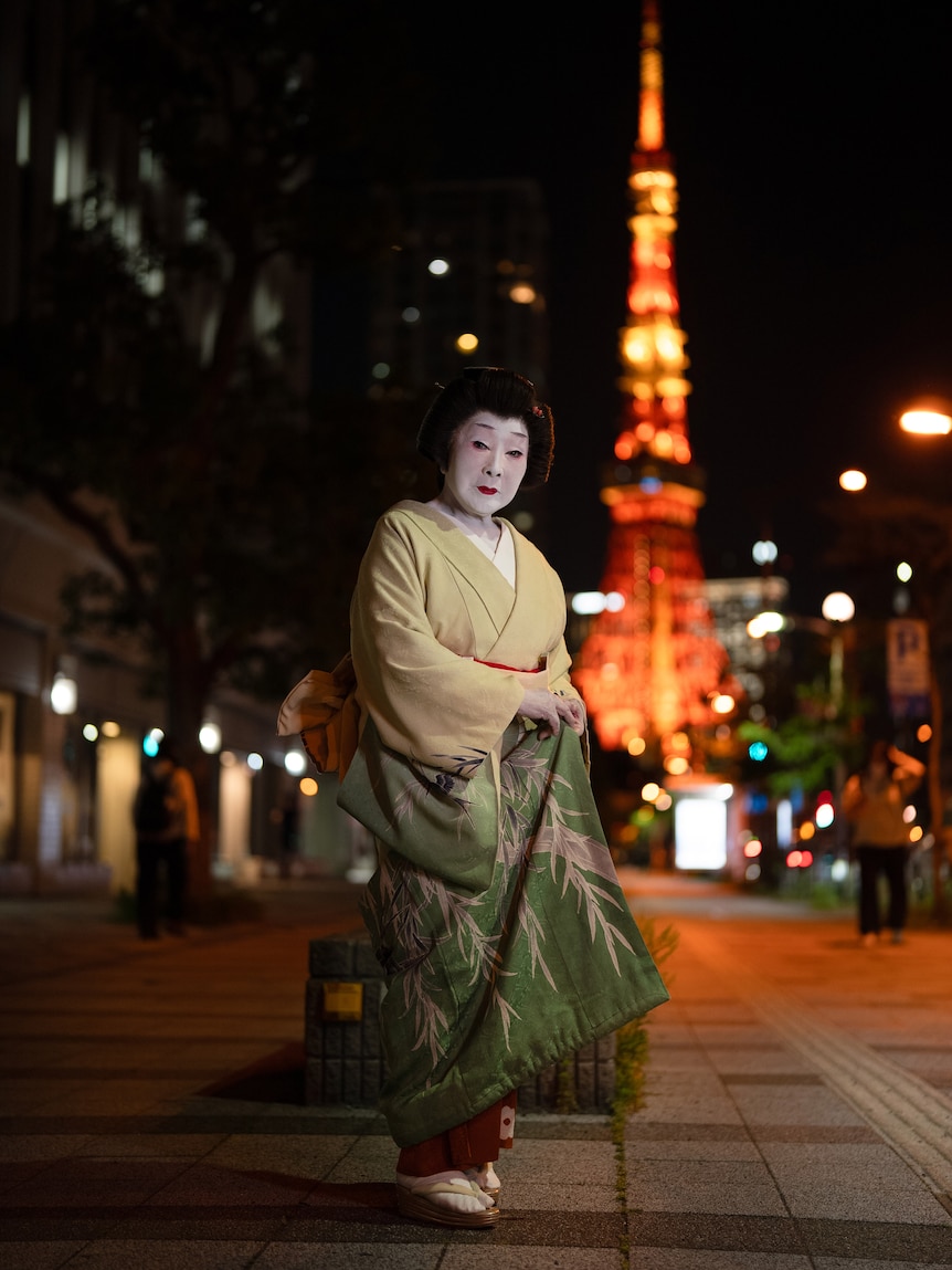 A woman in geisha dress stands in front of Tokyo Tower at night.