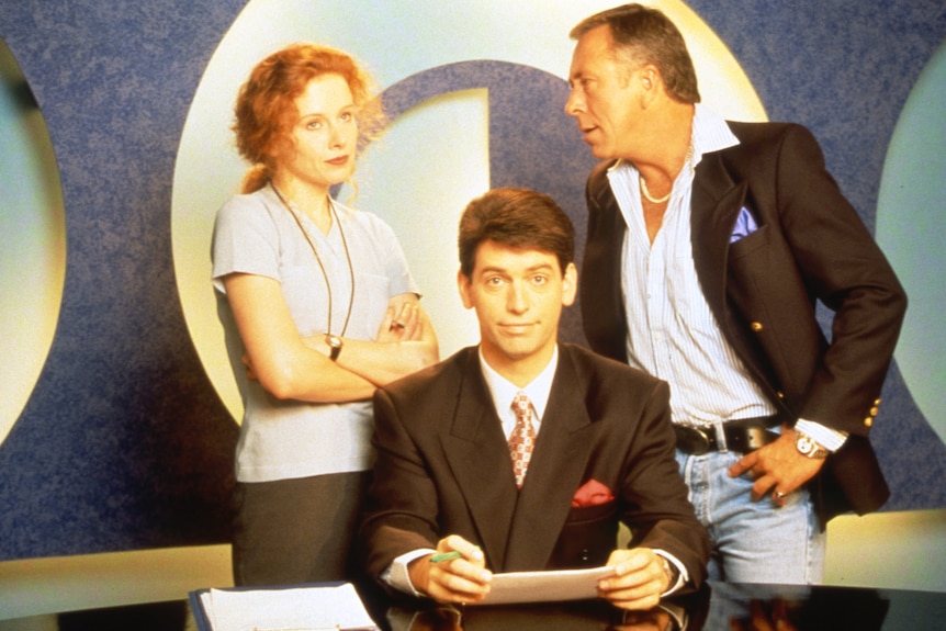 A TV still of Steve Bisley berating an exasperated Alison Whyte, Rob Sitch seated at news desk in front.