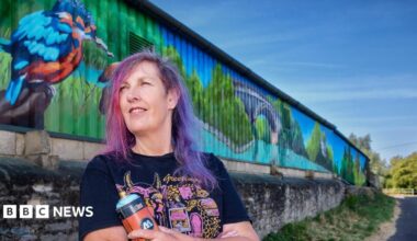 A woman stands in front of a colourful mural and holds a spray can. She has purple hair and glances off to the side