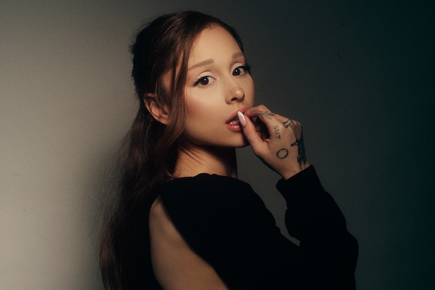 Ariana Grande with tattoos visible, holding a hand to her lips, side profile with skin showing.