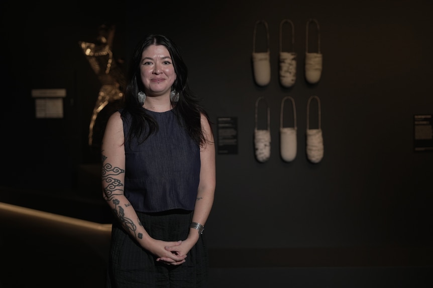 Jenna Mayilema Lee, an Indigenous woman in her early 30s, stands, hands clasped, in front of six woven bags on a gallery wall.