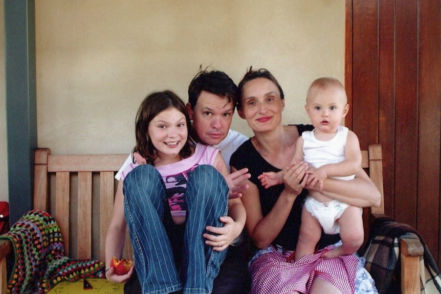 A man and woman seated on a wooden outdoor bench. A baby sits on the knee of mother, young girl on her father's lap