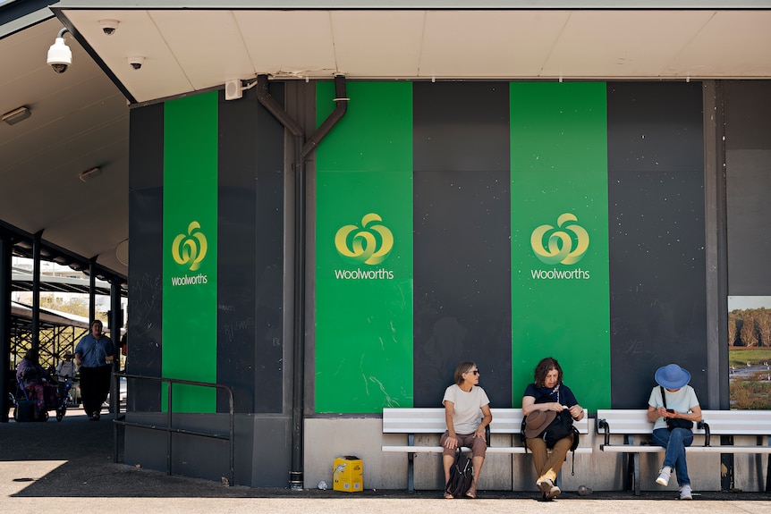 Three seated people waiting on a bench at a bus stop, the green Woolworths logo is on the wall behind them.