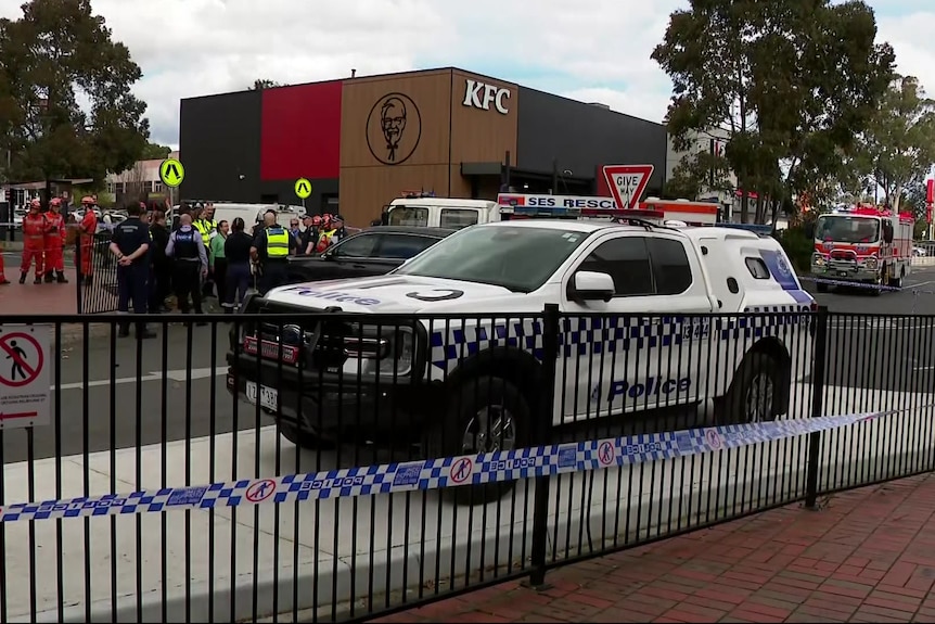 A large number of police and SES officers stand near a police car parked behind blue and white checked police tape.