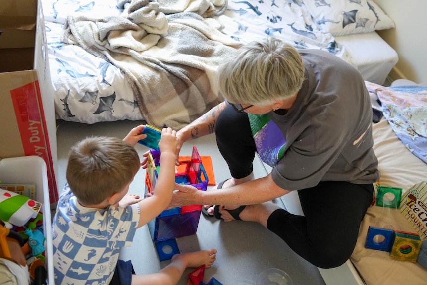 An aunt sits on the floor with a child with toys sprawled around them. They play with building blocks.