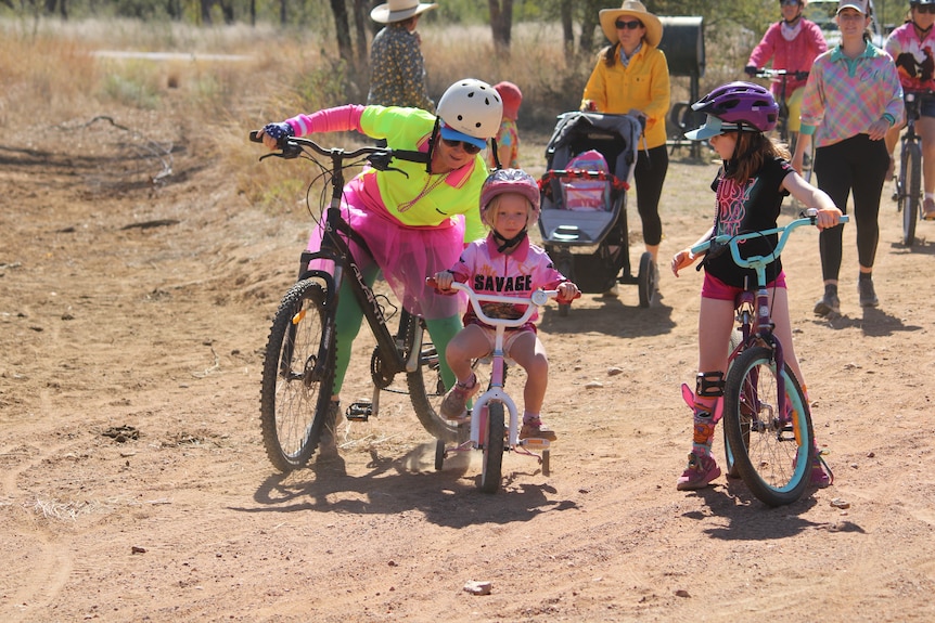 An adult helping push a young girl on a bike with training wheels during a race.