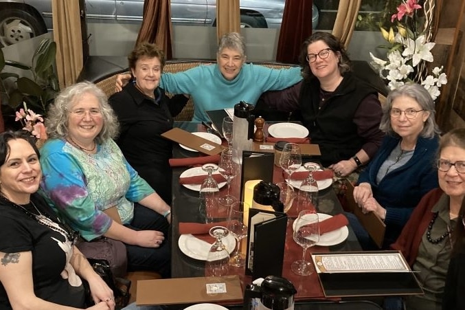 A group of women out for dinner. The woman in the centre has a blue jumper on and her arms around the others.